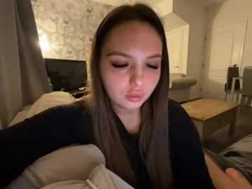 Video of a young woman with long brown hair and closed eyes, lying in bed with a blanket covering her chest, in a cozy, dimly lit bedroom with white walls and a wooden coffee table.