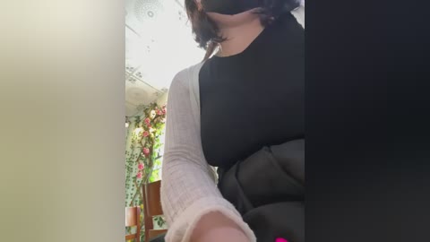 A video of a person wearing a black face mask, a black puffer jacket, and gray long sleeves, with a blurred background featuring greenery and a hanging floral arrangement.