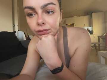 Video of a young woman with light skin, brown eyes, and short brown hair, lying on a bed, wearing a gray tank top and a black smartwatch, looking contemplative. Background shows a cozy, dimly-lit bedroom with a wooden dresser.