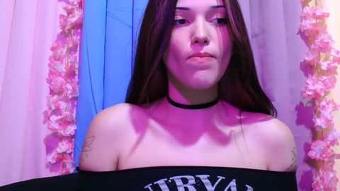 Video of a young woman with long brown hair, fair skin, wearing a black off-the-shoulder top with \"Nirvana\" text, black choker, and floral backdrop; purple lighting.