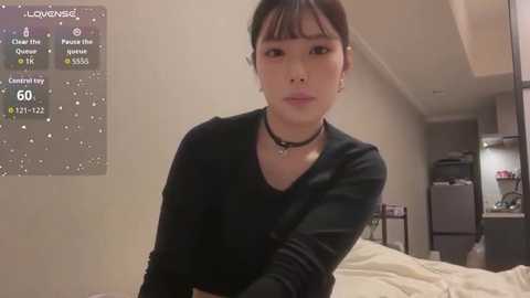 Video of a young Asian woman with fair skin and dark hair in a black long-sleeve top, sitting in a modern, minimalist bedroom with a digital screen displaying \"lovenss\" and \"center toy\" icons.
