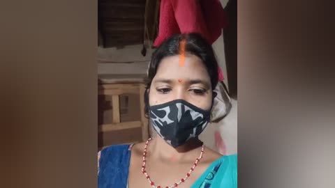 Video of a young woman with dark hair, wearing a blue sari, black mask with white skull design, red forehead mark, and orange dot. Background includes a wooden chair and wall.