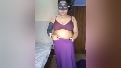 Video of a pregnant woman with medium skin tone, wearing a burgundy bra and purple maxi skirt, black mask, and arm cast, standing in a bedroom with a bed and wooden door.