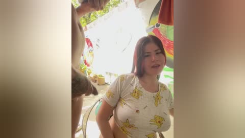 Video of a curvy woman with long brown hair in a white floral top, standing outdoors in a sunny, colorful garden setting.