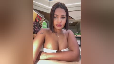 Video of a light-skinned woman with straight brown hair, wearing a white strapless top, sitting in a modern cafe with colorful fruit-themed wall art in the background.