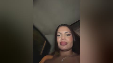 Video of a young woman with medium brown skin, long dark hair, and full lips, wearing minimal makeup, standing in a dimly lit room with blurred background.