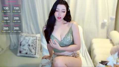 A video of an Asian woman with long black hair, wearing a glittery green bra and gold shorts, sitting on a white couch in a room with white curtains.