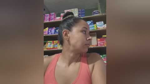 Media: Video of a Latina woman with dark hair in a bun, wearing a pink tank top, standing in a brightly lit convenience store with colorful snack shelves in the background.