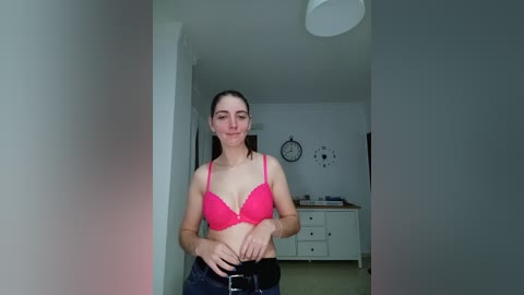 Media: A video of a young woman with light skin, dark hair, and a slim build, wearing a pink bra and black pants, standing in a minimalist room with white walls and a white dresser.
