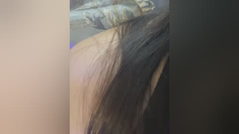 Video of a person's shoulder, showing long, straight, dark brown hair cascading down the left side. The background is blurred, with a hint of a light-colored blanket. The image is slightly out of focus, emphasizing the texture of the hair.