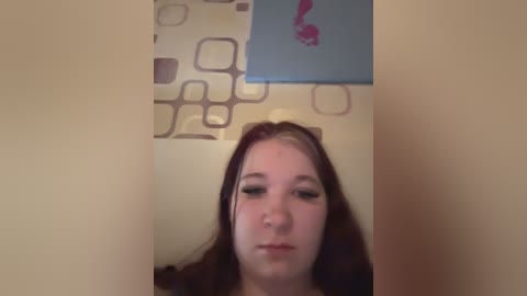 Video of a young woman with long brown hair, fair skin, and light makeup, standing against a beige wall with a light blue decorative panel.