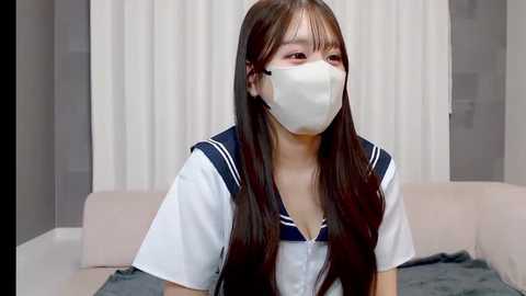 Video of a young East Asian woman with long, straight brown hair, wearing a white surgical mask, navy blue sailor-style school uniform, and sitting on a beige couch against a white curtain background.