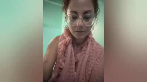 Video of a middle-aged woman with curly red hair and glasses, wearing a pink crocheted scarf and a sleeveless top, against a blurred green wall background.