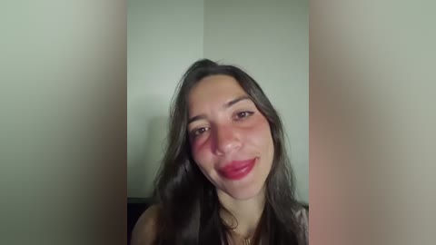 Video of a young woman with long, dark brown hair, fair skin, and full lips smiling. She wears a black top, and the background features a plain, light-colored wall.