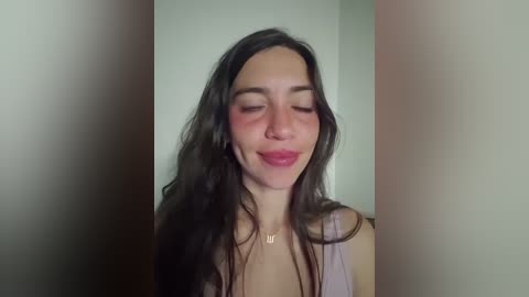 Video of a smiling woman with long dark hair, wearing a light pink tank top, and a gold necklace. Background is a plain, muted beige wall.