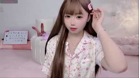 Video of a young Asian woman with long brown hair, wearing a floral shirt, adjusting her hair. Background includes a pink bedspread and a stuffed animal.