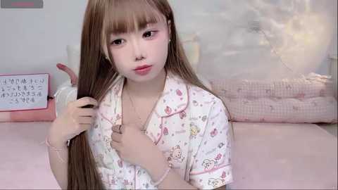 Video of a young Asian woman with long brown hair and bangs, wearing a pink pajama shirt with cartoon characters, sitting on a pink bed with a white wall and a pink lamp.