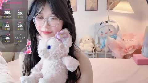 A video of a young Asian woman with long black hair, glasses, and a soft smile, holding a plush white bunny, surrounded by pastel decor in a cozy room.
