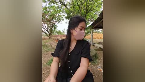Video of a young woman with long black hair, wearing a black dress, face mask, and sitting outdoors in a rural setting with trees and a small wooden structure.