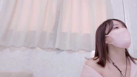 A video of an East Asian woman with straight, shoulder-length brown hair, wearing a pink face mask, a light pink off-shoulder top, and a black necklace, standing indoors with soft, white, sheer curtains in the background.