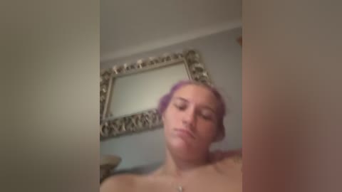 Video of a topless woman with light skin and purple hair, leaning forward with a neutral expression, in a dimly lit room with an ornate mirror and a beige wall.