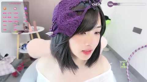 A video of an East Asian woman with short black hair, wearing a purple lace bonnet, in a well-lit room. Background includes a white wall, a small table, and a green chair.