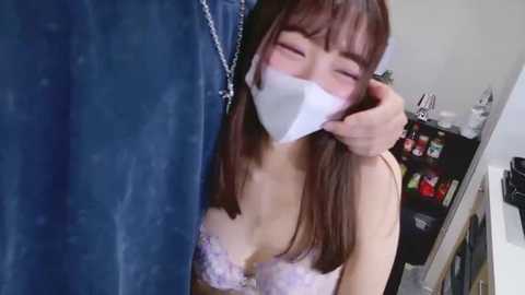 Video of a young Asian woman with long brown hair, wearing a pink lace bra, and a white face mask, smiling while being held by a man in a blue denim jacket.