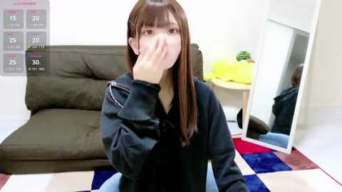 A video of an Asian woman with long brown hair, wearing a black sweater, covering her mouth in a dark living room with a green couch, yellow pillow, and a colorful rug.