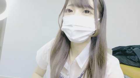 Video of an East Asian woman with long, straight, brown hair, wearing a white mask, white collared shirt, and plaid tie, against a plain, white background.