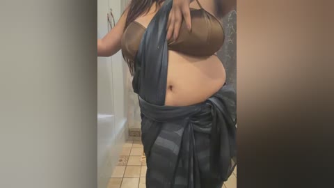 Media: Video of a woman with long dark hair, wearing a brown bra and a dark blue sari, standing in a tiled bathroom. She covers her chest with her hand.