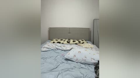 Video of a messy, unmade bed in a small, dimly lit room. The bed features a light gray headboard, and the bedding is a mix of blue, white, and patterned sheets.