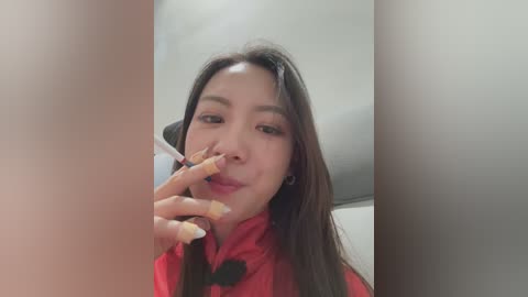 A video of a young East Asian woman with long dark hair, wearing a red dress, holding a cigarette to her lips, smiling, indoors.