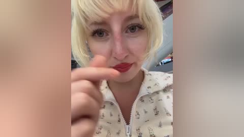 Video of a young woman with short, platinum blonde hair, pale skin, and red lipstick, wearing a light-colored hoodie with bunny print, biting her finger, blurry background.