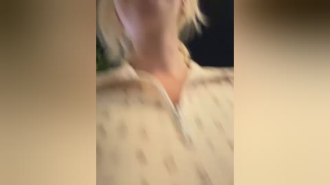 A video of a blonde woman with shoulder-length hair, wearing a white blouse with subtle horizontal stripes, focusing on her neck and upper chest. The background is blurred, suggesting an indoor setting.