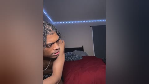 Media: Video of a Black man with dreadlocks, wearing a necklace, lying on a bed with red sheets, looking sad, in a dimly lit room with blue LED strip lighting on the ceiling.