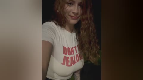 Video of a smiling woman with long curly auburn hair, wearing a white t-shirt with red \"Don't Jealous Me\" text, indoors against a dark background.