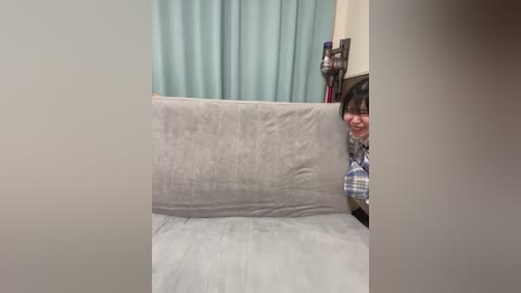 Video of a young Asian woman with glasses and short black hair peeking out from behind a gray couch. She wears a blue and white plaid shirt. Background includes teal curtains and a bookshelf.