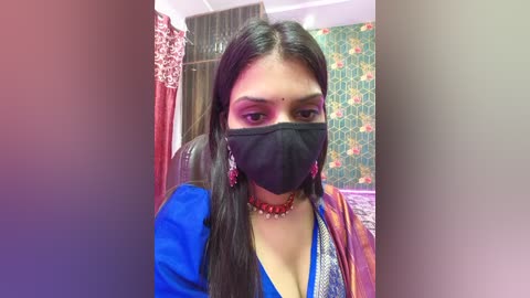 Video of a woman with long black hair, wearing a blue and red traditional saree, black face mask, and red earrings, indoors with colorful wallpaper and decorative curtains in the background.