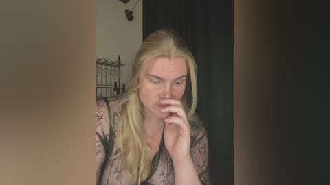 Video of a blonde woman with fair skin, wearing a sheer lace top, sitting pensively, fingers to lips, in a dimly lit room with a black curtain.