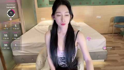 Video of a young Asian woman with long black hair, wearing a lace-up corset, sitting on a beige chair in a modern bedroom.