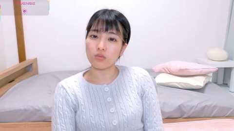 Video of a young East Asian woman with short black hair, wearing a light grey, knitted cardigan, sitting on a bed in a minimalist, white-walled room.