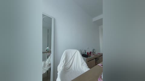 Video of a minimalist, sparsely furnished bedroom with white walls, a wooden desk, and a large mirror reflecting part of the room. A white towel is draped over a chair.