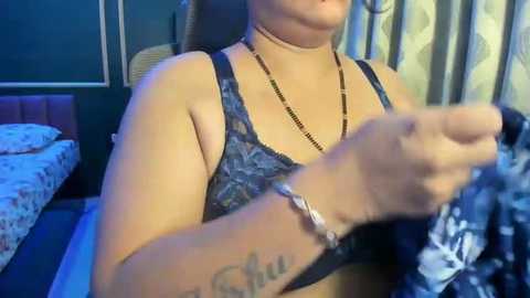 Video of a light-skinned woman in a black floral bra, holding a patterned fabric, wearing a black beaded necklace and wristband. Background shows a bed with floral sheets, dark curtains, and a wooden headboard.