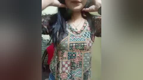 Video of a woman with long, dark hair, wearing a colorful, patterned dress with geometric designs, posing indoors. She stands against a blurred background with muted tones.