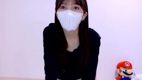 Video of an Asian woman with long, straight brown hair, wearing a white face mask, black top, and white gloves, standing against a plain white wall. Mario plush toy in red and blue to the right.