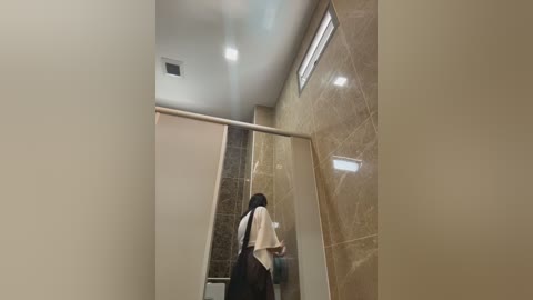 Video of a modern, beige-tiled bathroom with a woman with long black hair, wearing a white top and dark pants, drying her hair with a towel, standing near a glass shower door.