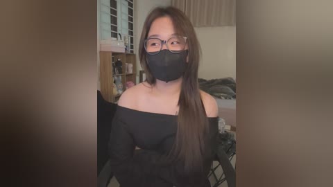 Video of an Asian woman with long black hair, wearing glasses, a black mask, and off-shoulder black top, standing in a cluttered bedroom with beige walls, a bed, and a shelf.