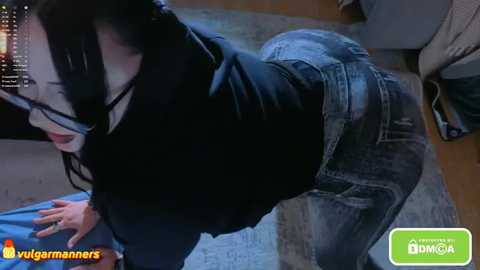 A video captures a woman in a black top and jeans, leaning over a table, her long dark hair cascading. The background features a grey sofa and a blue cushion. The scene is dimly lit, with a watermark of \"vulgarmanns\" and \"DOMICIA.\