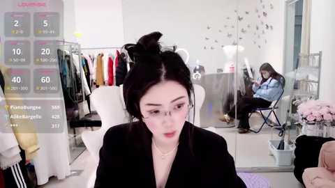 Video of a chic, minimalist clothing store with a young woman in glasses and black hair tied up, sitting at a table. Background features white walls, mannequins, and a woman seated in the corner.