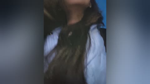 A close-up video of a person's neck and upper chest, wearing a black fur-trimmed white jacket over a white shirt, with a blurred background of blue and gray.